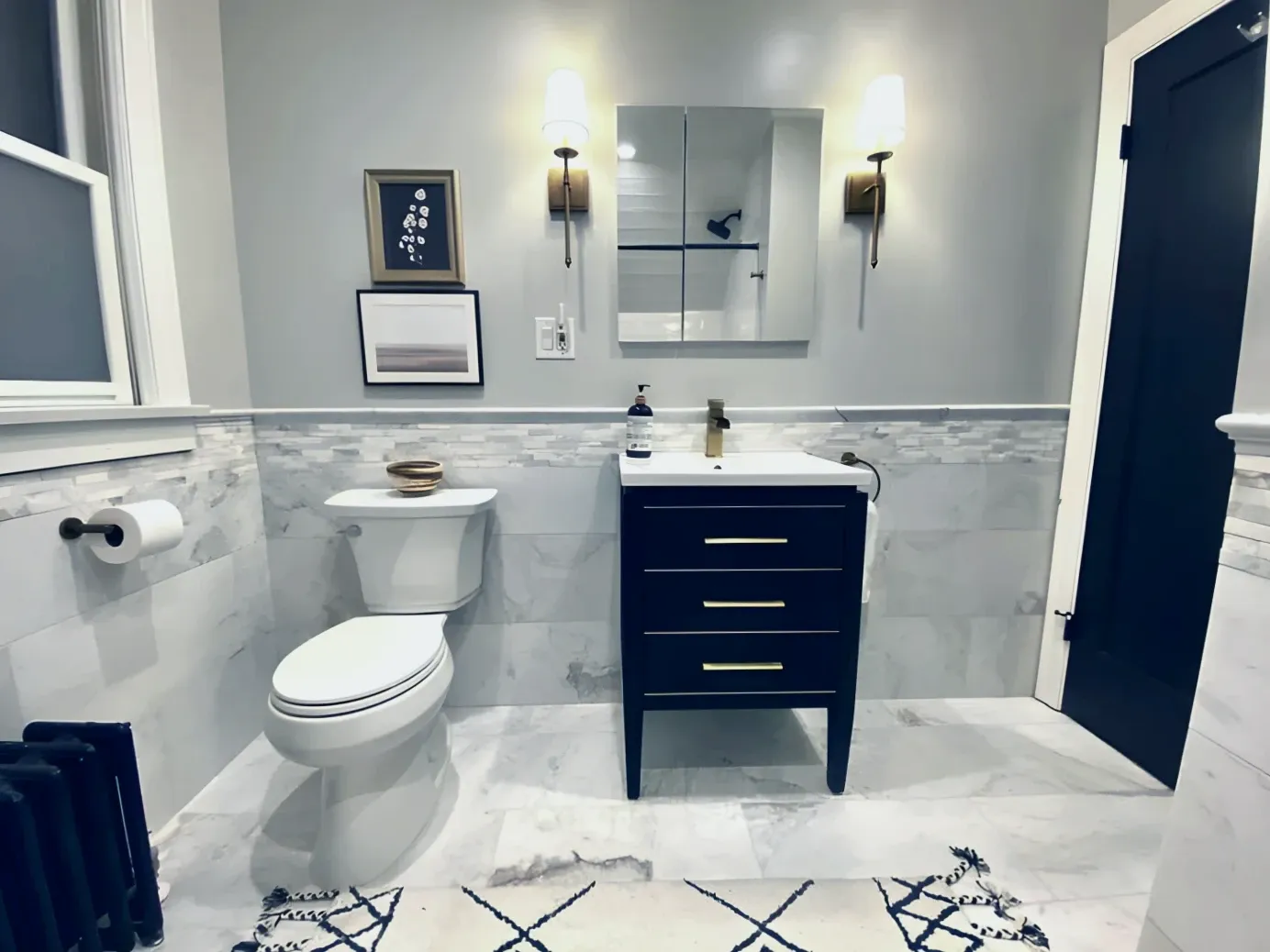 Clean white modern bathroom with attractive rug, art, and black set of drawers. The clean mirror is flanked by two modern, torch-like lamps.