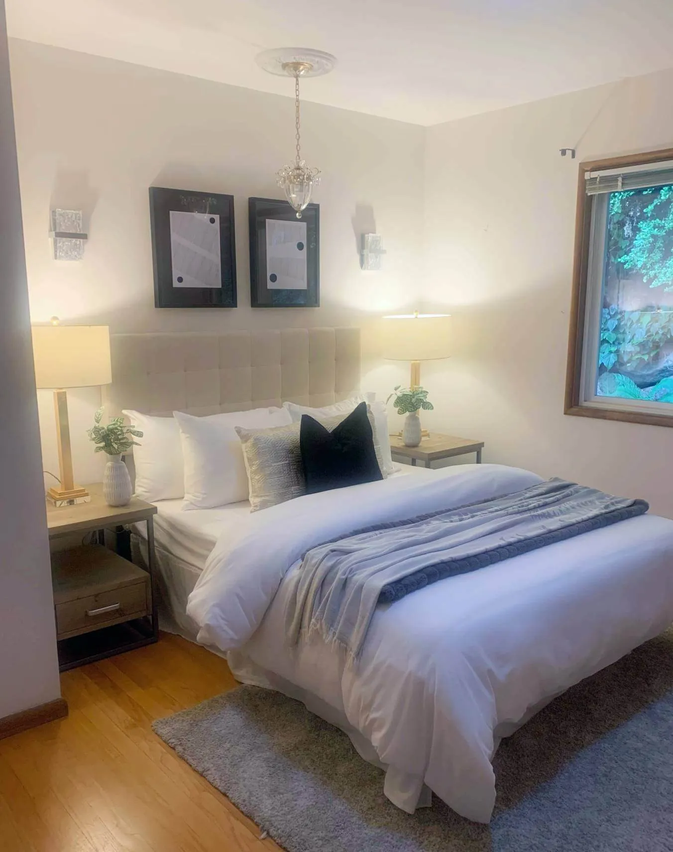 Small, modern bedroom with decorative pillows and blankets. Mirrored bedside tables and tall lamps on either side of the queen bed.