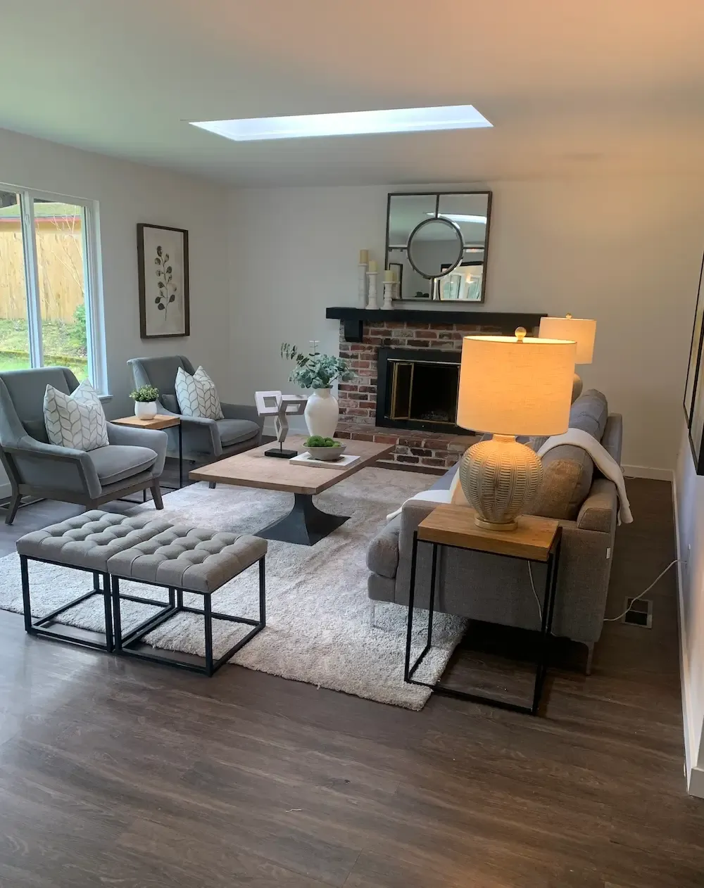 A cozy living room with modern design featuring a gray sofa, two armchairs, and a tufted bench. A coffee table is in the center, a fireplace with a mirror and decorations above it, and lamps on side tables. Large windows and a skylight bring in natural light.