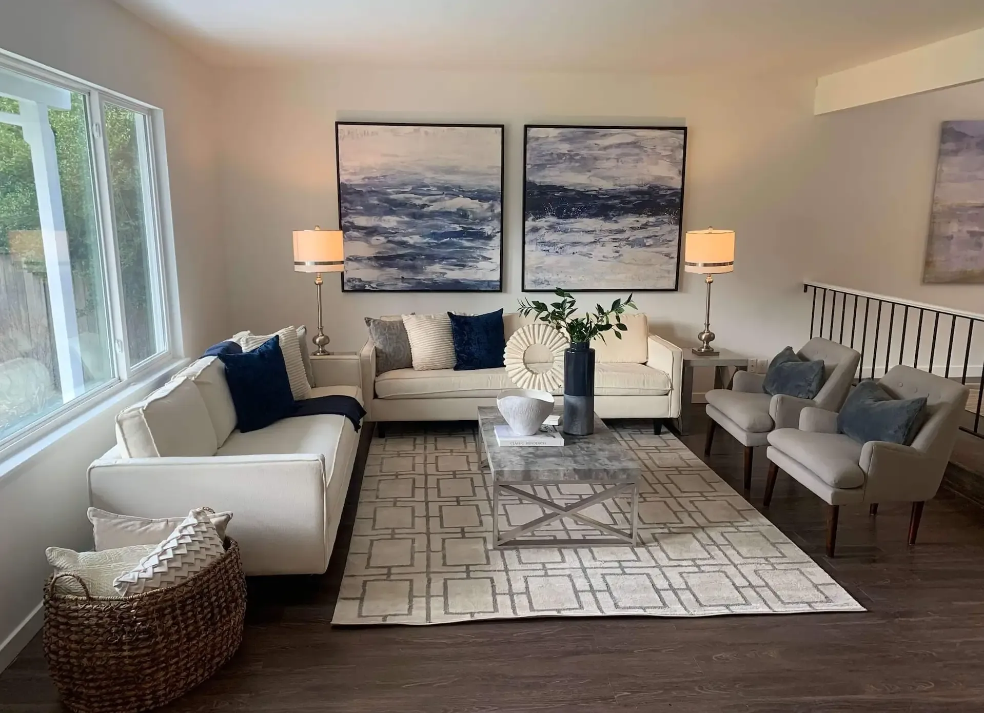 A modern living room with two cream sofas and two gray armchairs surrounding a glass coffee table on a patterned rug. Two large blue and white abstract paintings hang above the sofas. Lamps with white shades and a basket of pillows complete the decor.