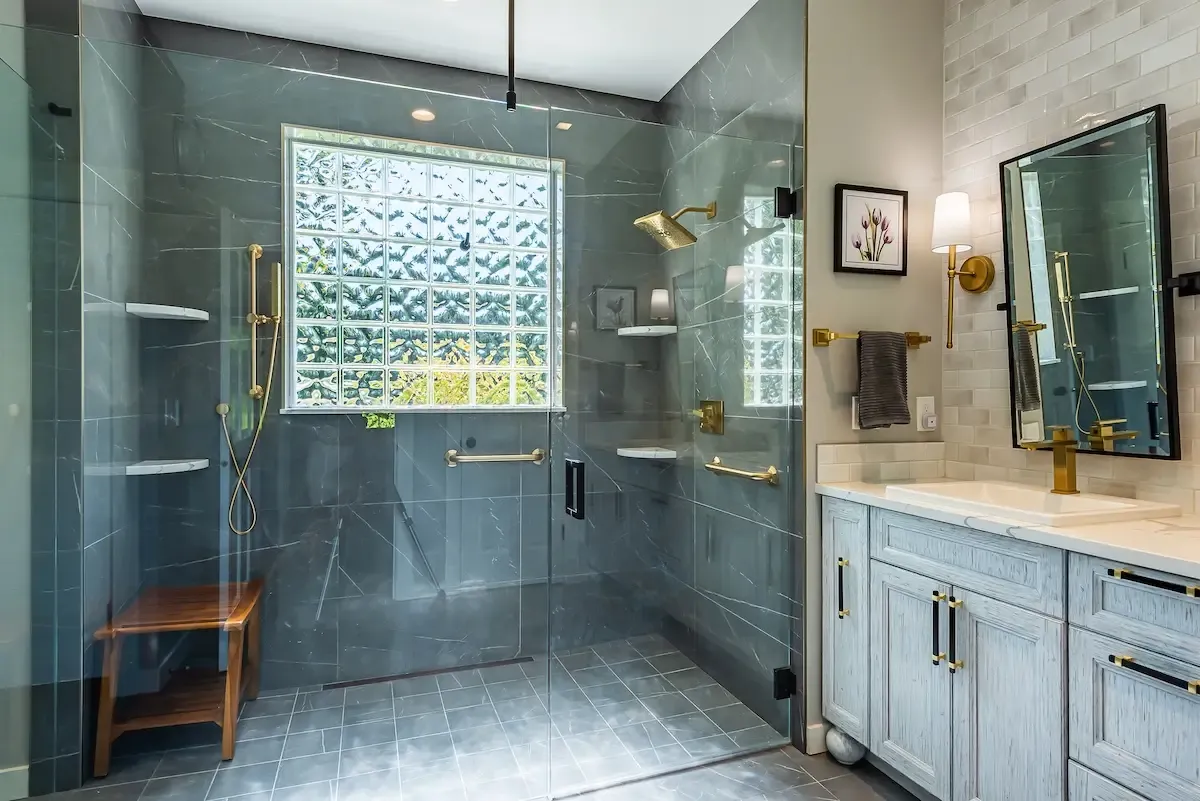 A modern bathroom featuring a large glass shower enclosure with gray tiled walls, a wooden bench, and dual showerheads. A vanity with double sinks, gold fixtures, and a mirror is adjacent to the shower. A large glass-block window lets in natural light.