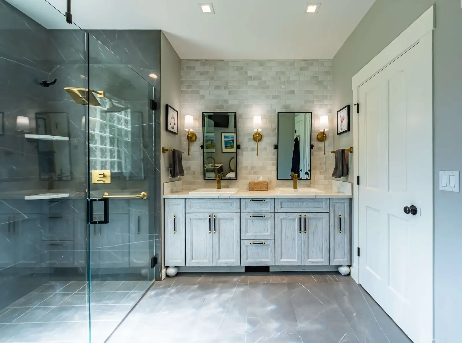 A modern bathroom featuring a gray double vanity with two mirrors and wall-mounted lights. To the left, a glass-enclosed shower has a gold showerhead and fixtures. The walls have a mix of gray tile and white accents, and the floor is tiled in gray.