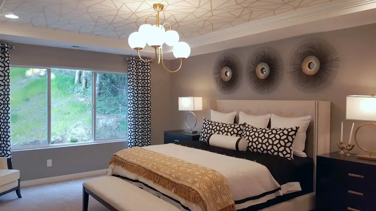 A modern bedroom with a large window, patterned curtains, and a bed featuring geometric-patterned bedding. Above the bed are three decorative wall art pieces. A stylish chandelier hangs from the ceiling, and there are two nightstands with lamps on each side of the bed.