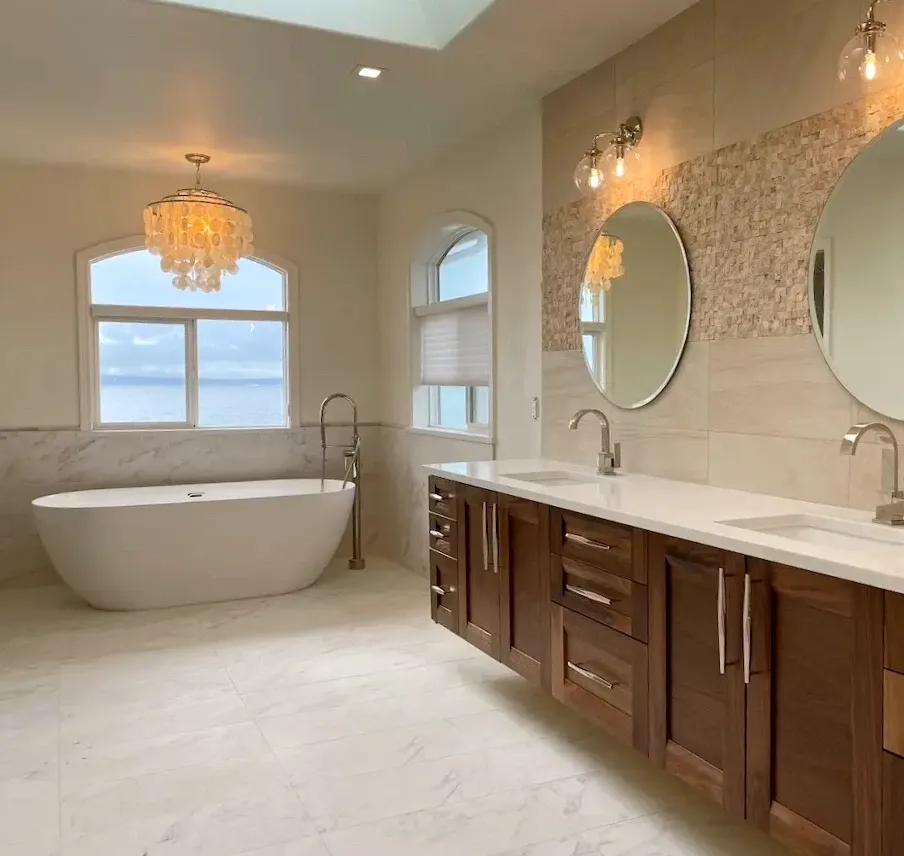 Spacious bathroom featuring a freestanding white bathtub, dual wooden vanities with white countertops and round mirrors, and modern light fixtures. The room has large windows with ocean views and a light-colored tiled floor. A chandelier hangs from the ceiling.