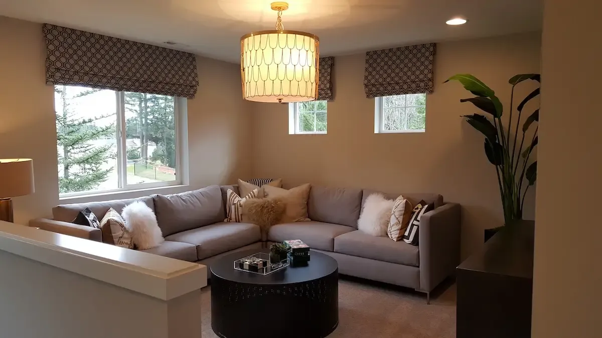 A cozy living room with a large sectional sofa adorned with pillows, a round coffee table with decorative items, and a tall potted plant. The room has a chandelier and multiple windows with patterned valances, allowing natural light to filter in.