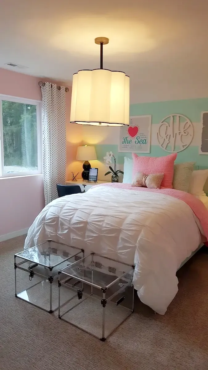 A cozy bedroom with a large window on the left, a bed adorned with white and pink pillows and a comforter. Two transparent cubes act as a bench at the foot of the bed. A desk with a computer, lamp, and decorative items is in front of the window. Modern decor on the walls.