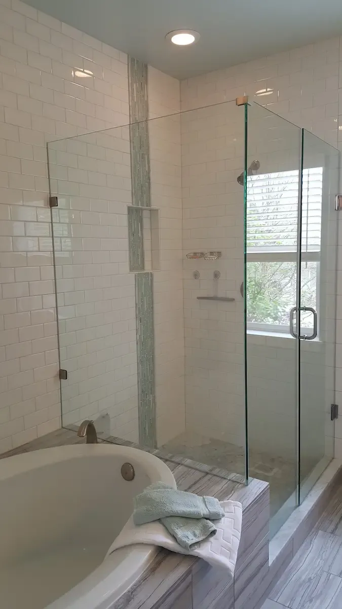 A modern bathroom featuring a glass-enclosed walk-in shower with white subway tile walls and a vertical accent strip. A soaking tub with folded green towels on its edge is adjacent. Sunlight filters through a window near the shower.