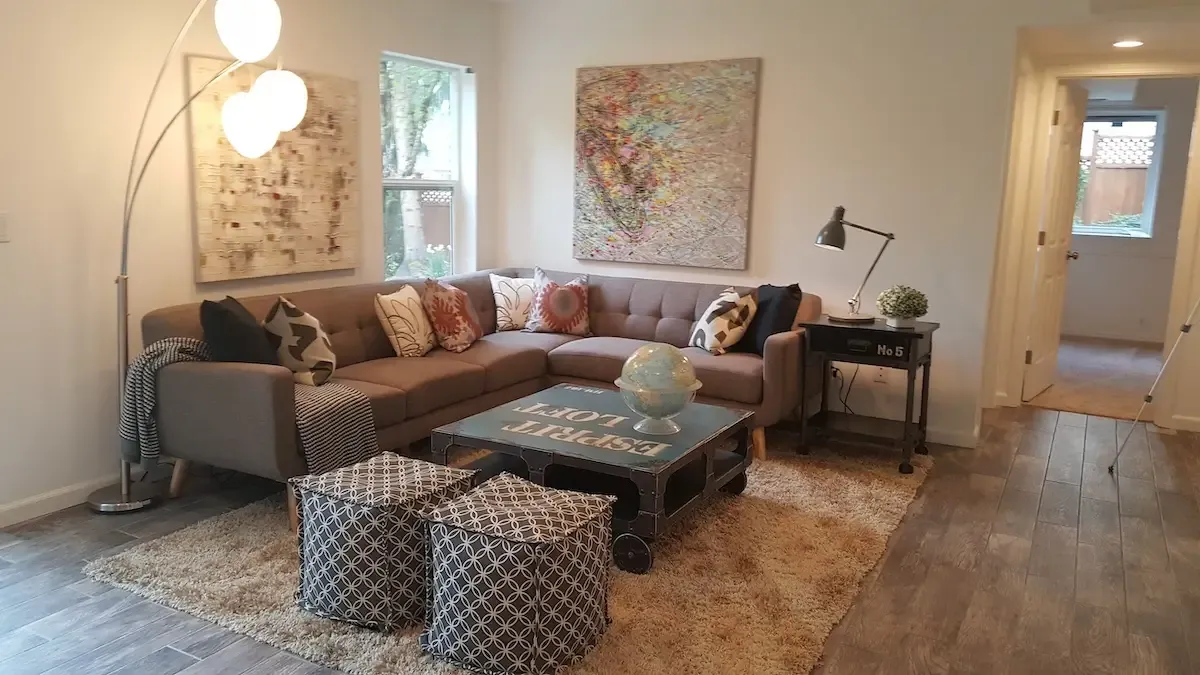 A cozy living room with modern decor. It features a gray L-shaped couch adorned with various throw pillows, a coffee table with a globe on top, two patterned ottomans, abstract art on the walls, a floor lamp, and a wooden floor with a beige rug.