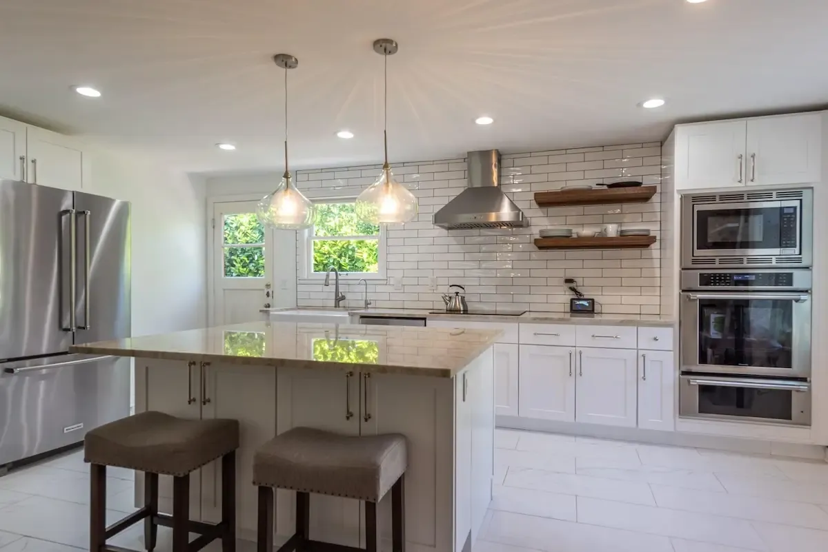 Gleaming white kitchen with brick-like white tiles on the wall, decorative curved hanging lamps, and beige stone countertops.