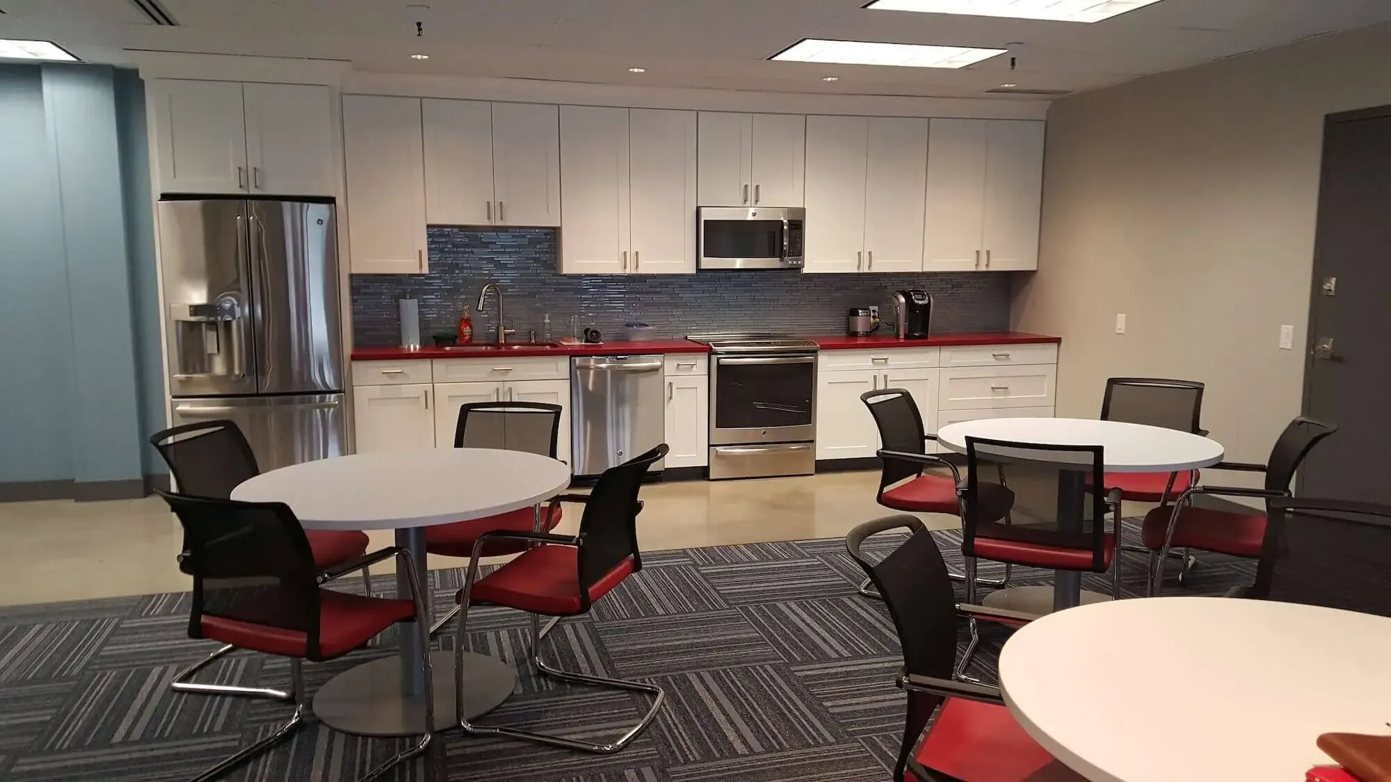 Tidy office break room space with three tables and fetching black and red chairs. Strong gray and white contrasts in the kitchen.