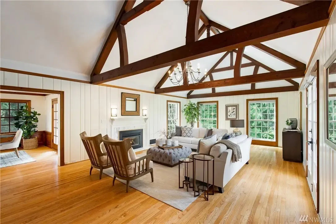 A spacious living room with wooden flooring and a high ceiling featuring exposed wooden beams. The room is furnished with a white sectional sofa, two wooden chairs, a tufted ottoman, and a fireplace.