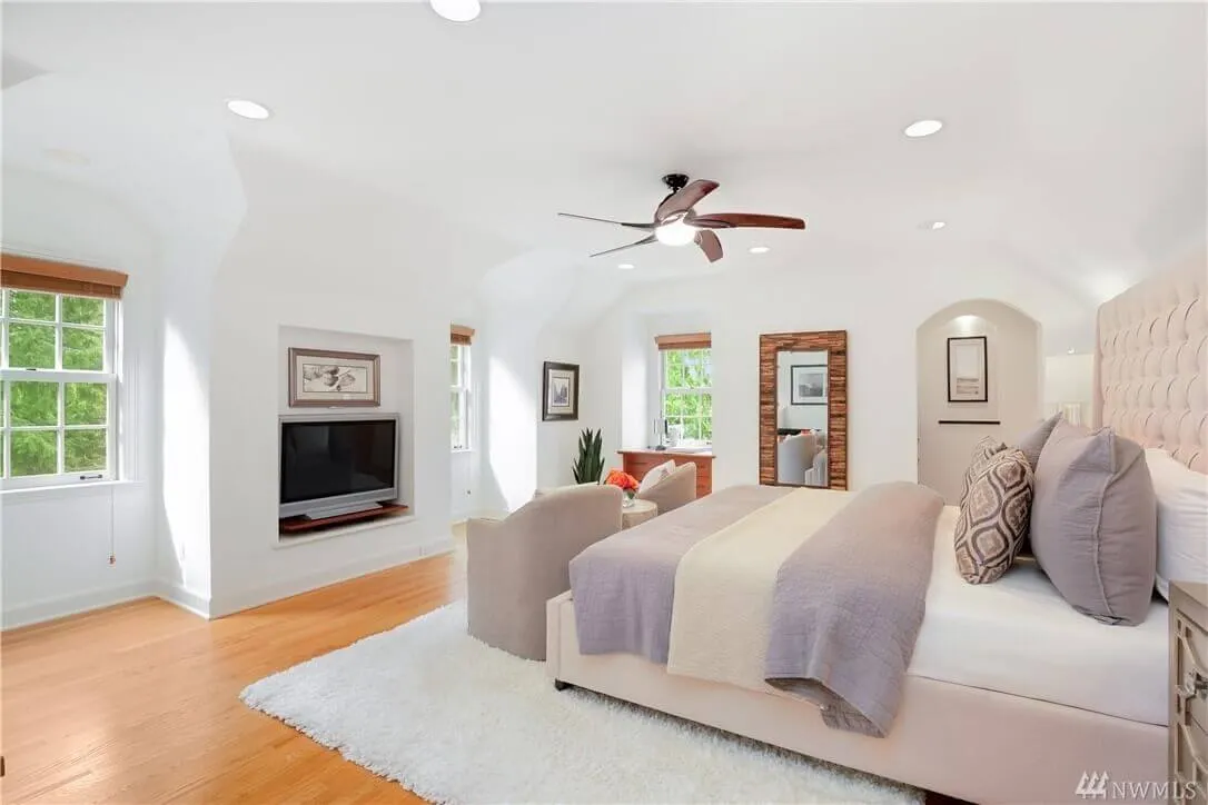 A bright and spacious bedroom with white walls and hardwood flooring. The room features a large bed with gray and white bedding, a ceiling fan, two windows with wooden blinds, and a wall-mounted TV. A plush beige armchair sits at the foot of the bed.