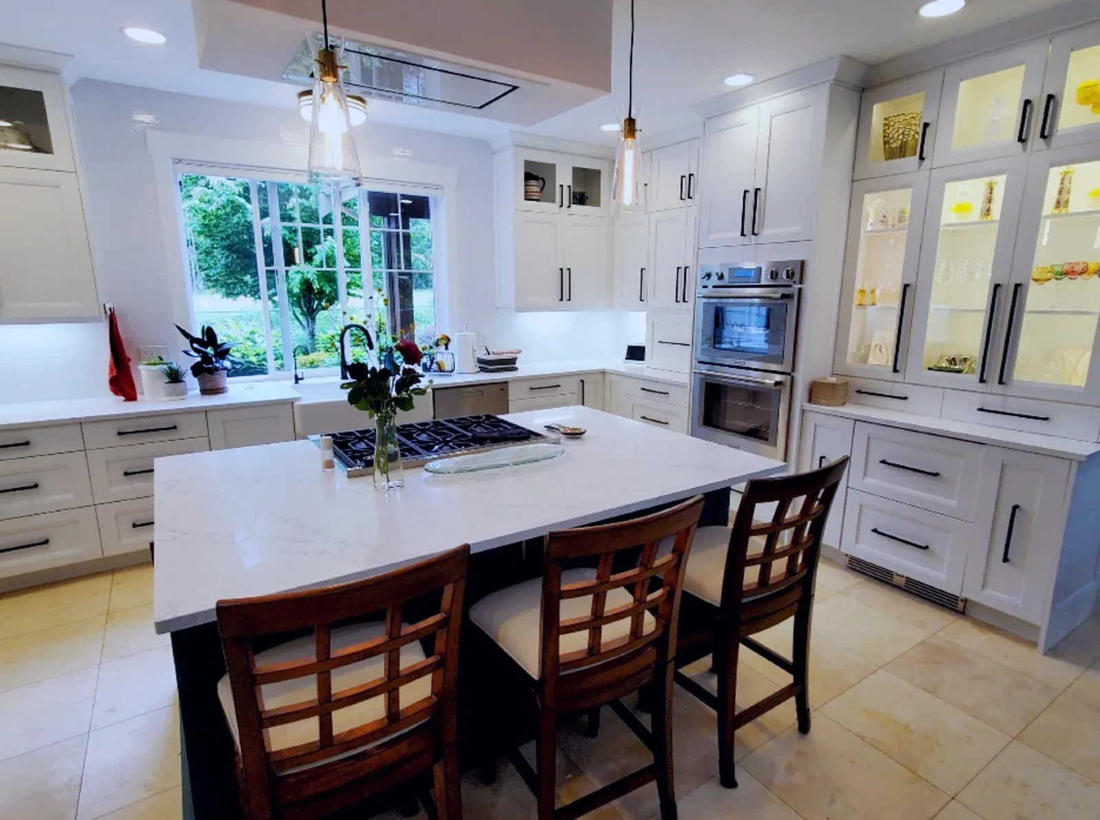 a sleek, modern kitchen with all white cabinetry. Some cabinets have glass panes and lights inside which illuminate decorative china. A set of three chairs sit by the center island. The countertops are white quartz.